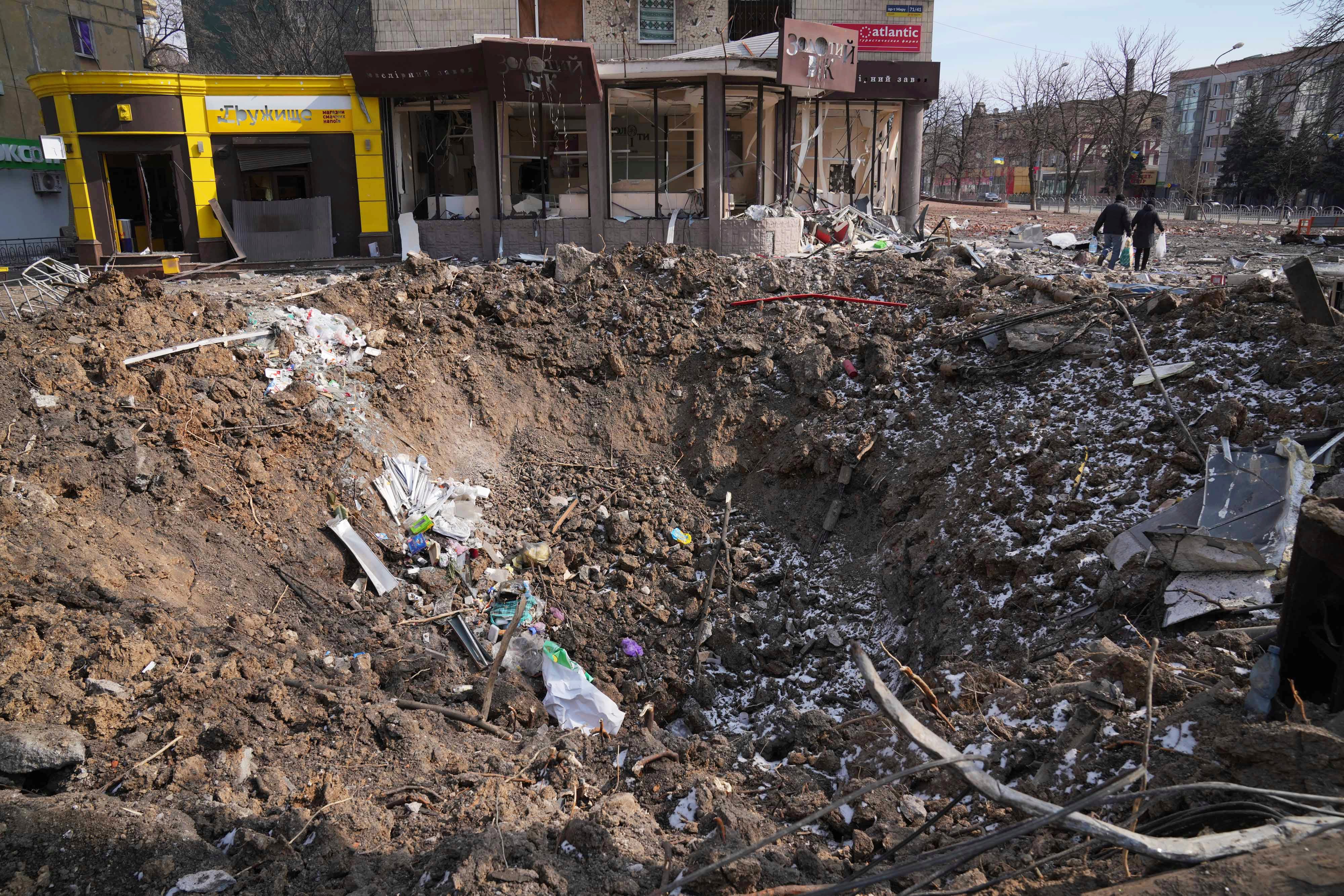 People walk past a crater from an explosion on Mira Avenue (Avenue of Peace) in Mariupol, Ukraine, Sunday, March 13, 2022. The surrounded southern city of Mariupol, where the Russian war on Ukraine has produced some of the greatest human suffering, remained cut off despite earlier talks on creating aid or evacuation convoys. (AP Photo/Evgeniy Maloletka)